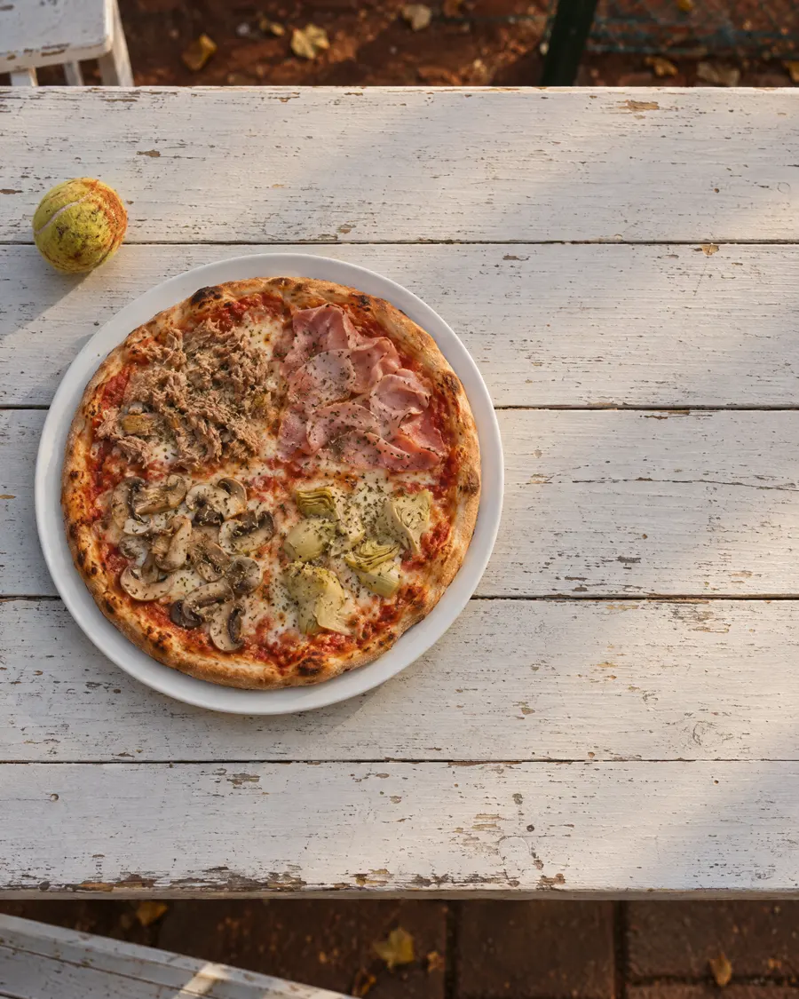 Pizza Quattro Stagioni von oben auf einem weißen Teller, daneben ein Tennisball